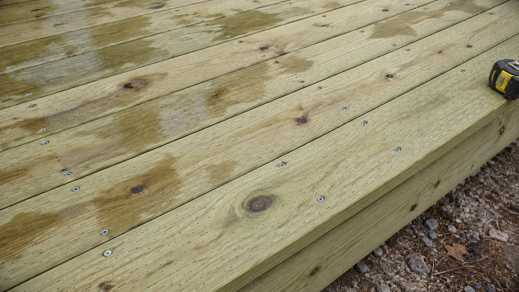 Freshly laid pressure treated pine decking with knots, a green treatment tint and rows of countersunk screw heads, a tape measure at the edge